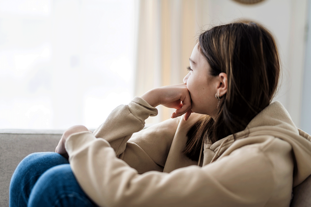 a young adult woman sits with her legs up and rests her chin on her first while looking out the window wondering about the connection between trauma and addiction
